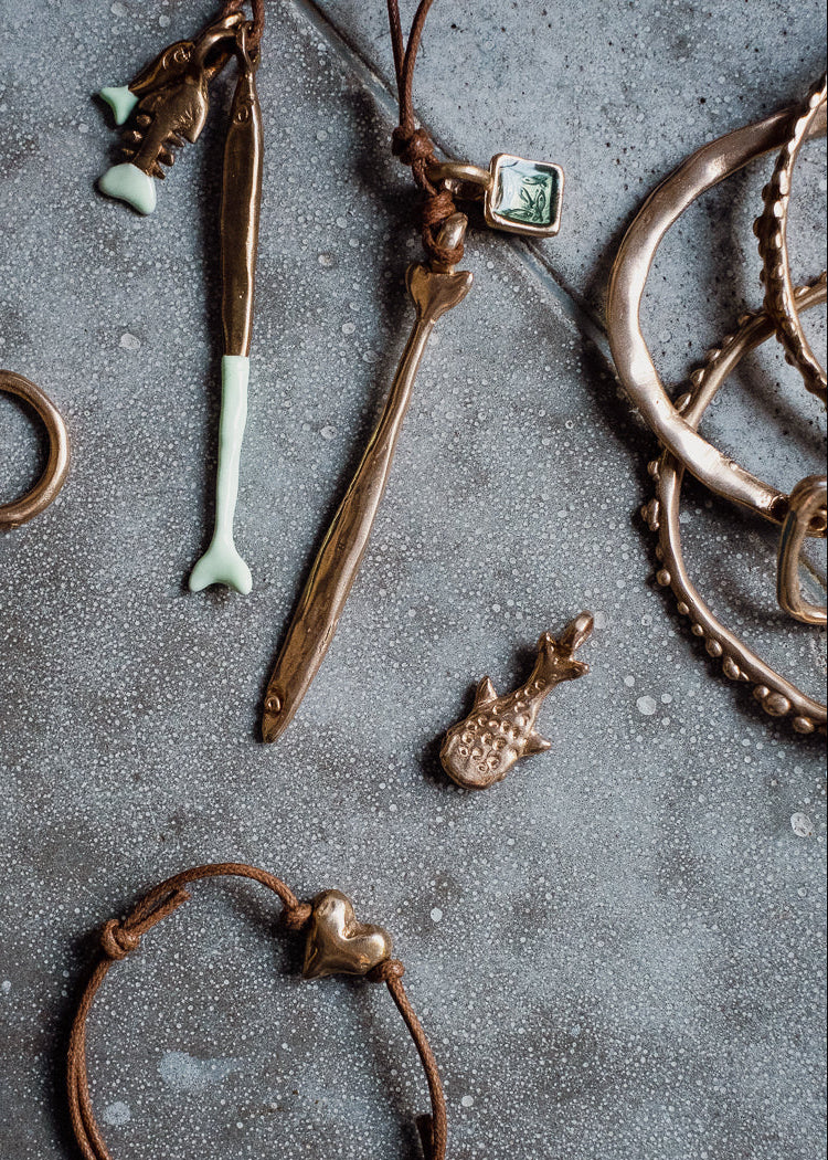 A styled flat-lay of various handmade jewelry pieces, including a brown leather bracelet with a small metallic charm, a bronze-toned arrow pendant, a small green stone charm, and multiple layered metal rings, arranged on a textured gray concrete surface.